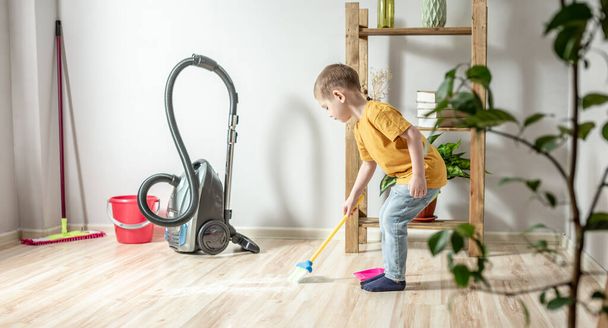 A cute little boy is sweeping the trash from the floor with a broom to a dustpan. Child helps the parents in cleaning the house - Foto, Bild