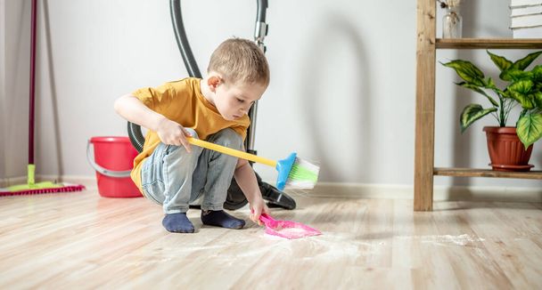 A cute little boy is sweeping the trash from the floor with a broom to a dustpan. Child helps the parents in cleaning the house - Foto, afbeelding