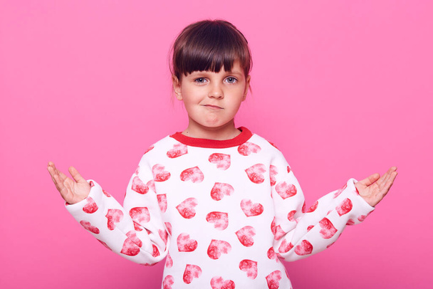 Little dark haired female kid posing with spreading hands aside, looking at camera with frowning face, wearing casual attire, , isolated over pink background. - Photo, Image