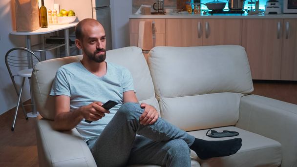 Уставший мужчина сидит перед телевизором и смотрит развлекательные фильмы - Фото, изображение
