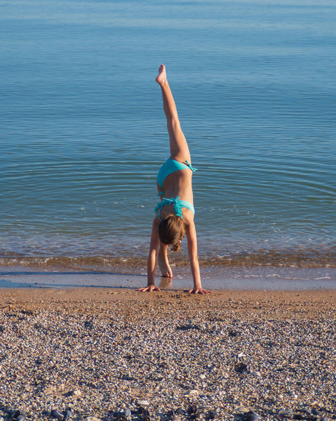 Teinityttö voimistelee rannalla veden äärellä. Tyttö sinisessä uimapuvussa hiekkarannalla lähellä taivaansinistä merta. Ulkoilu kunto ja terveellinen elämäntapa. - Valokuva, kuva
