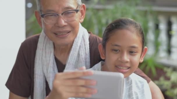 おじいさんの横に座っているアジア系の愛らしい孫娘は、自宅のスマートフォンカメラでオンラインでビデオ通話から彼女の友人に挨拶するために手を振って話すのを楽しんでいます。家族の絆. - 映像、動画