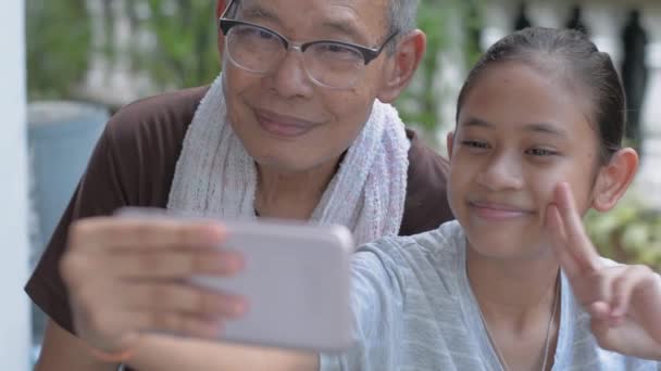 Šťastná rozkošná asijská dívka se svým starším dědečkem vychutnat legrační pózování selfie fotografie a natáčení videa se sociálními médii on-line z mobilního smartphonu v jejich domě. - Záběry, video