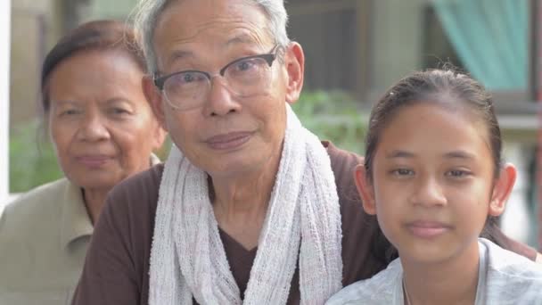 Portret van gelukkige bejaarde gepensioneerde grootouders met schattige kleindochter die samen zitten en glimlachen terwijl ze naar de camera kijken tijdens hun verblijf thuis. Samenzijn in twee generaties. Relatie tussen gezin en gezin. - Video