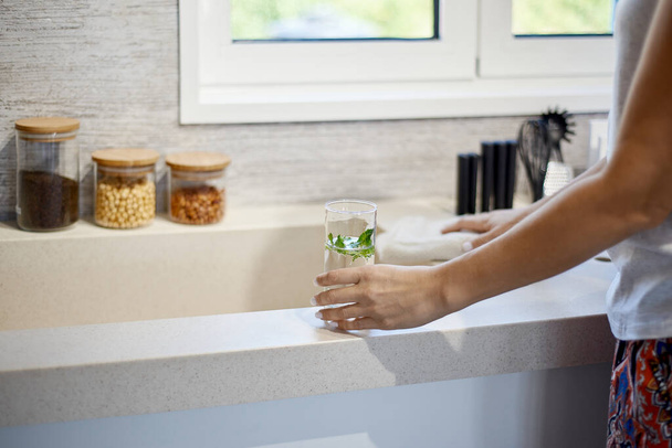Egy nő egy pohár vizet tart a mosogató mellett a konyhában. - Fotó, kép