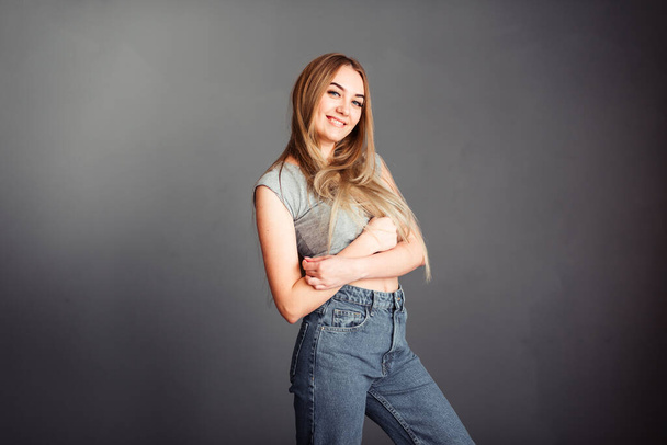 Foto retrato de una hermosa chica sonriente sobre un fondo gris en jeans y una blusa con largo cabello rubio hermoso. De pie justo delante de la cámara. - Foto, imagen