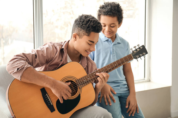 Έφηβος με τον μικρό του αδερφό να παίζει κιθάρα στο σπίτι. - Φωτογραφία, εικόνα