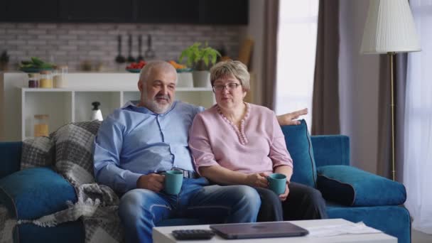 starší muž a žena komunikují v obývacím pokoji svého bytu, důchodci sedí spolu na gauči a pijí čaj - Záběry, video