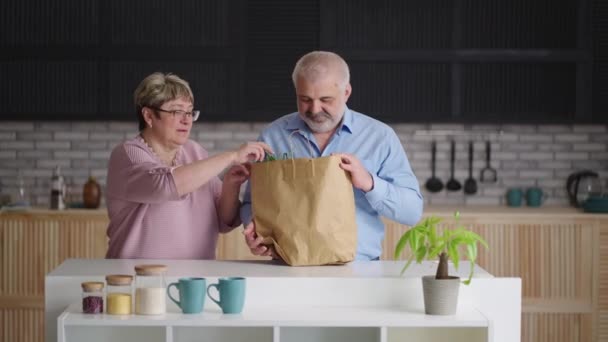 starší muž a žena vybalují nákupy z potravin nebo trhu, dávají čerstvou zeleninu na kuchyňský stůl z papírového sáčku - Záběry, video