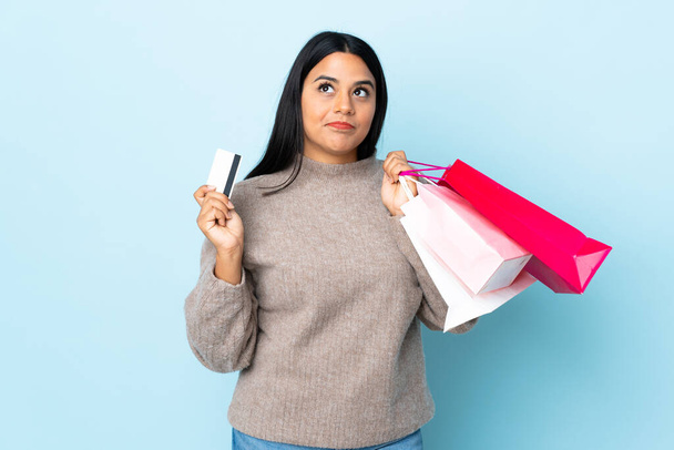 Young latin woman woman isolated on blue background holding shopping bags and a credit card and thinking - Fotoğraf, Görsel