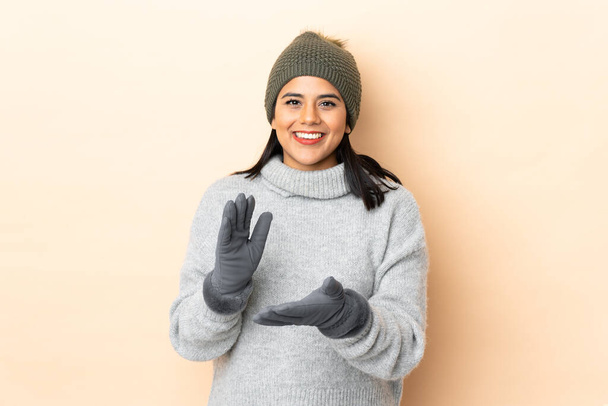 Νεαρή Κολομβιανή κοπέλα με χειμερινό καπέλο απομονωμένη σε μπεζ φόντο χειροκροτεί - Φωτογραφία, εικόνα