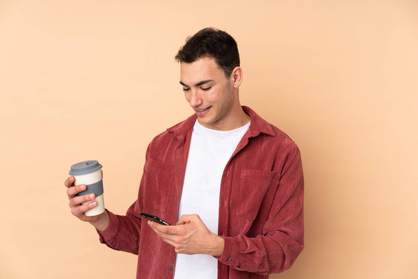 Young caucasian handsome man isolated on beige background holding coffee to take away and a mobile - Photo, Image