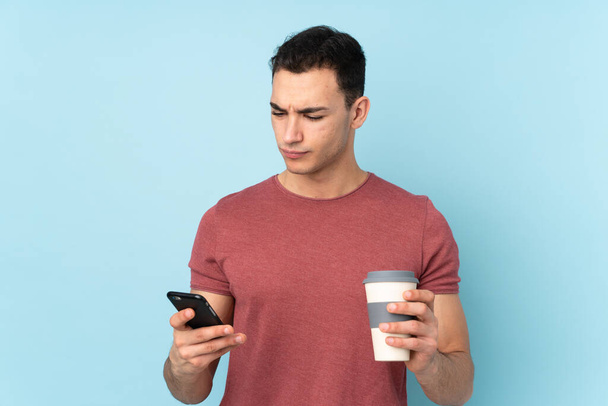 Jonge blanke knappe man geïsoleerd op blauwe achtergrond met koffie om mee te nemen en een mobiele - Foto, afbeelding