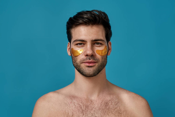 Portrait of brunette caucasian guy with golden gel under eye patches looking at camera, posing isolated over blue background - Photo, image