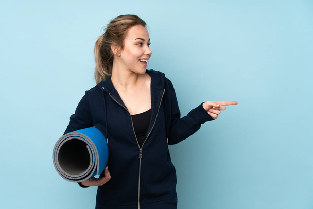 Teenager Russian girl holding mat isolated on blue background pointing finger to the side and presenting a product - Photo, Image