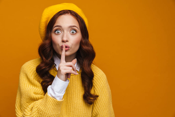 Happy young woman with long red hair in sweater and beret standing over yellow wall background, showing silence gesture - 写真・画像