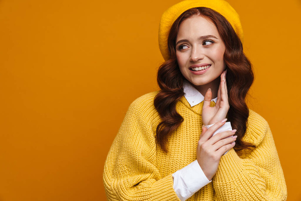 Happy young woman with long red hair in sweater and beret standing over yellow wall background, lookng aside at copy space - Fotó, kép