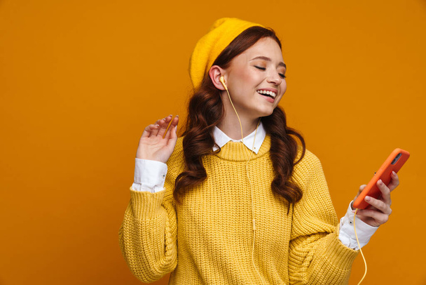 Glückliche junge Frau mit langen roten Haaren in Pullover und Baskenmütze, die über gelbem Wandhintergrund steht und ihr Handy in der Hand hält - Foto, Bild