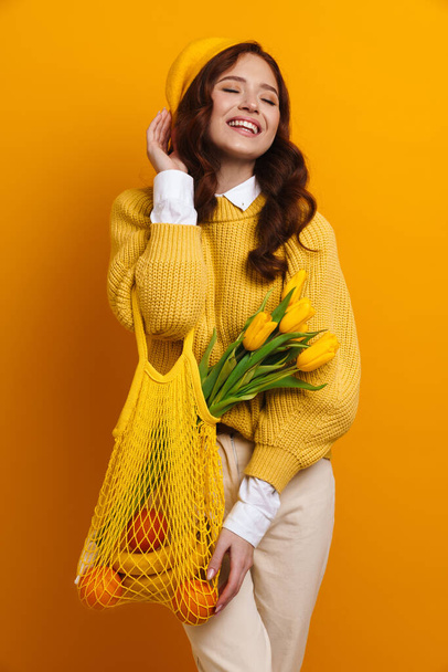 Smiling young woman with long red hair in sweater and beret standing holding tulips bouquet carrying bag with fruits over yellow wall background - Photo, Image
