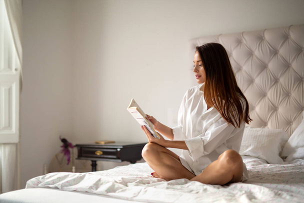 Odpoczynek, komfort, wypoczynek i koncepcja ludzi. Sexy szczęśliwy kobieta czytanie książki w łóżku w domu - Zdjęcie, obraz