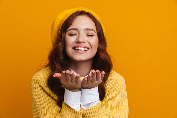 Happy young woman with long red hair in sweater and beret standing over yellow wall background, sending love kiss - Fotografie, Obrázek