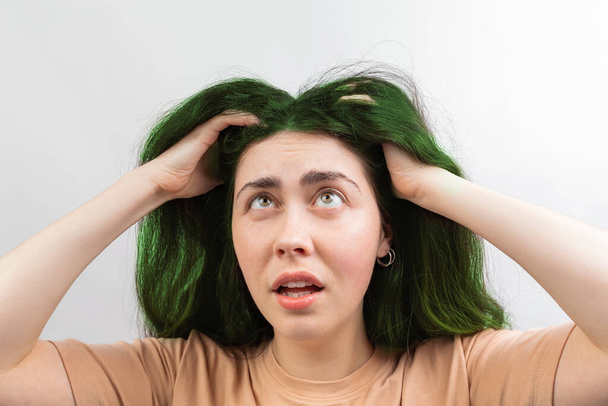 Hair coloring. A young Caucasian woman in a beige t-shirt, looking up at her long green hair in fright. White background. - Photo, Image