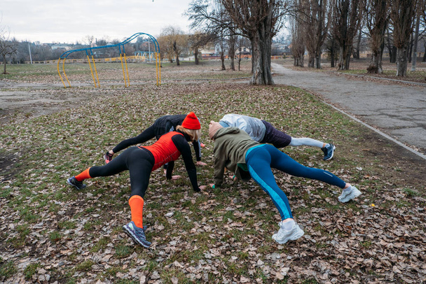 Групповые занятия по фитнесу на открытом воздухе. Social Distant Outdoor Workout Classes in public parks. Три женщины и мужчина тренируются вместе в общественном парке. Здоровье, благополучие и общественная концепция - Фото, изображение