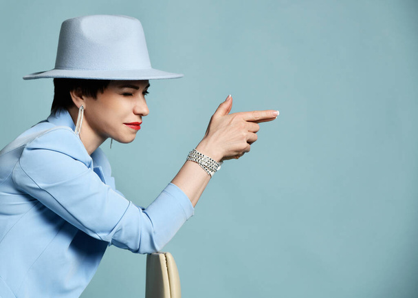 Cheerful brunette woman in blue business suit and wide-brimmed hat takes aim with her finger gun. Side view - Foto, afbeelding