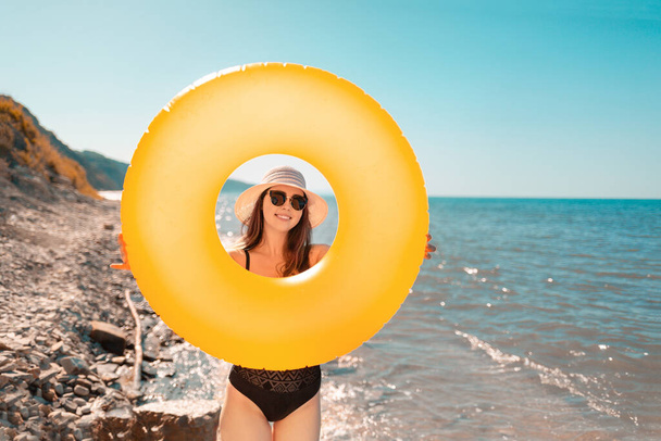 Una mujer joven bronceada sostiene un círculo inflable anaranjado y mira a través de él.En el fondo es una playa salvaje. El concepto de unas vacaciones de verano en el mar, viajar y nadar. - Foto, imagen
