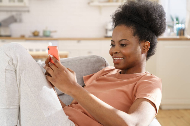 Happy african girl lying on sofa use smartphone texting in social media, reading sms with good news - Foto, Bild