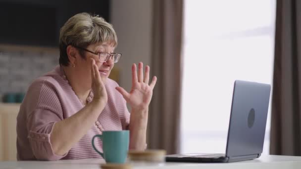 fröhliche ältere Dame kommuniziert mit Familie per Videochat im Laptop, ältere Frau sitzt am Wochenende in der heimischen Küche - Filmmaterial, Video