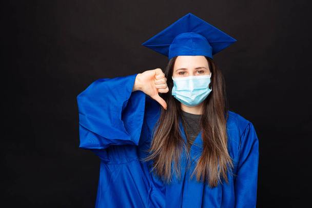 Foto di giovane donna laureata mostrando pollice verso il basso, antipatia e indossare maschera facciale. - Foto, immagini