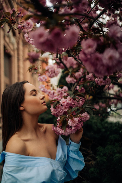 Young woman has enjoy and relaxes in park with blooming sakura trees against building background. Portrait. - Fotoğraf, Görsel