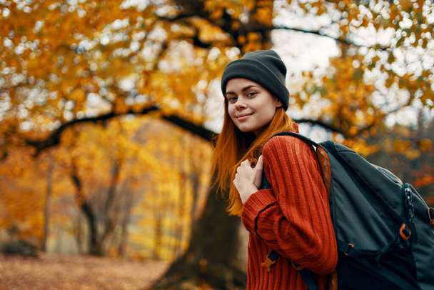 Porträt einer Frau mit Pullover und Rucksack auf dem Rücken in der Nähe der Bäume im Herbst im Wald - Foto, Bild
