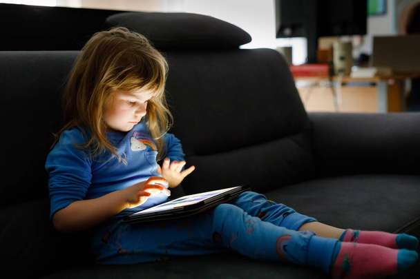 Χαριτωμένο κοριτσάκι παίζει με το tablet pc στο σπίτι. Υγιές μωρό αγγίζοντας pad με τα δάχτυλα, αναζητούν κινούμενα σχέδια και να διασκεδάσουν με εκπαιδευτικές εφαρμογές. Παιδί με πιτζάμες νυχτικό ακούω ακουστικό βιβλίο - Φωτογραφία, εικόνα