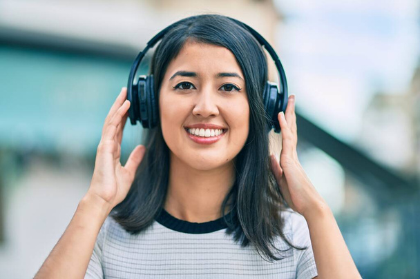 Jonge Spaanse vrouw die blij glimlacht en muziek luistert met een koptelefoon in de stad. - Foto, afbeelding