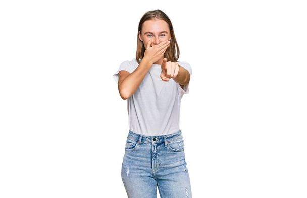 美しい若いブロンドの女性はカジュアルな白いtシャツを着てあなたを笑って、口の上に手でカメラに指を指して、恥の表情  - 写真・画像