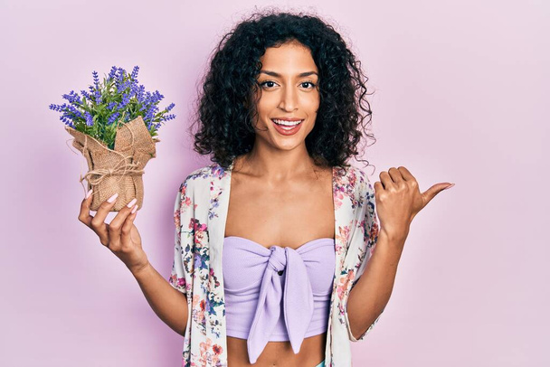 Nuori latino tyttö tilalla laventeli kasvi osoittaa peukalo ylös sivulle hymyillen onnellinen suu auki  - Valokuva, kuva