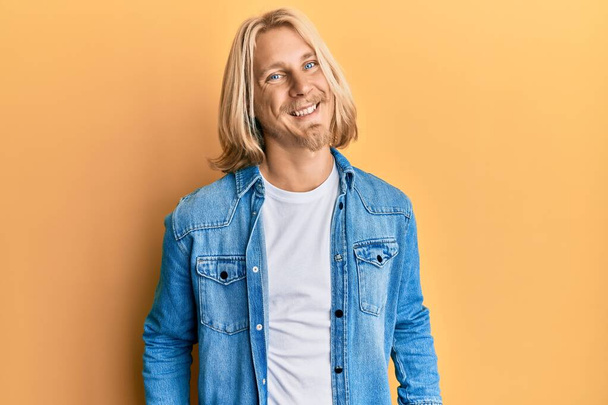 Homme caucasien aux cheveux longs blonds portant une veste en denim décontractée avec un sourire heureux et cool sur le visage. chanceux.  - Photo, image