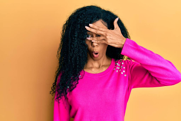 Middelbare leeftijd Afrikaanse Amerikaanse vrouw dragen casual kleding gluren in shock bedekken gezicht en ogen met de hand, kijken door vingers bang  - Foto, afbeelding