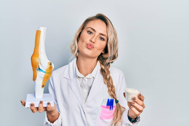 Krásná mladá blondýnka lékař žena drží anatomický model kolenního kloubu a bílkovin prášek při pohledu na fotoaparátu foukání polibek je krásné a sexy. výraz lásky.  - Fotografie, Obrázek