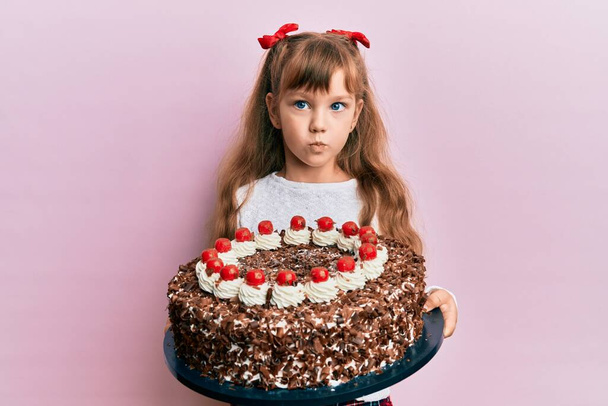 Ένα μικρό καυκάσιο κοριτσάκι γιορτάζει τα γενέθλιά του κρατώντας ένα μεγάλο κέικ σοκολάτας κοιτάζοντας την κάμερα να φυσάει ένα φιλί είναι υπέροχο και σέξι. έκφραση αγάπης.  - Φωτογραφία, εικόνα
