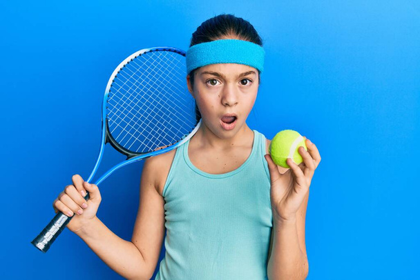 Beautiful brunette little girl playing tennis holding racket and ball in shock face, looking skeptical and sarcastic, surprised with open mouth  - Photo, Image