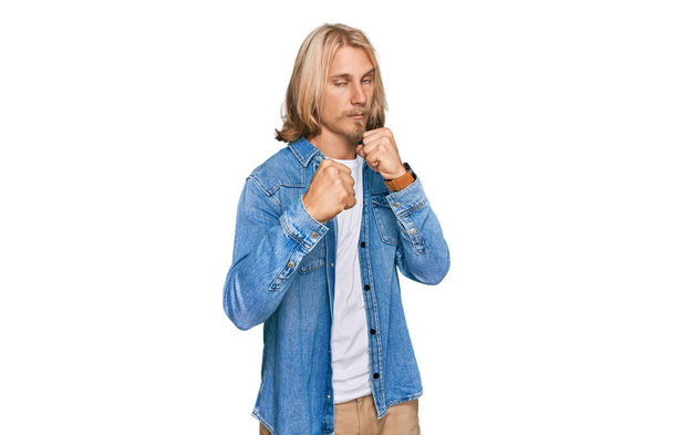 Caucasian man with blond long hair wearing casual denim jacket ready to fight with fist defense gesture, angry and upset face, afraid of problem  - Fotografie, Obrázek