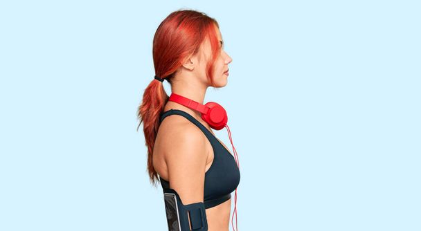 Jeune femme rousse portant des vêtements de gymnastique et utilisant des écouteurs regardant vers le côté, pose de profil relax avec visage naturel avec sourire confiant.  - Photo, image