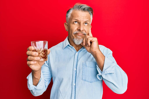 Μεσήλικας γκριζομάλλης άντρας που πίνει ένα ποτήρι ουίσκι σοβαρά σκεπτόμενος την ερώτηση με το χέρι στο πηγούνι, σκεπτόμενος την μπερδεμένη ιδέα  - Φωτογραφία, εικόνα