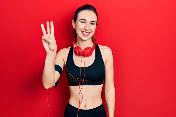 Молода іспаномовна дівчина носить спортивний одяг і використовує навушники, показуючи і вказуючи пальцями номер три, посміхаючись впевнено і щасливо
.  - Фото, зображення