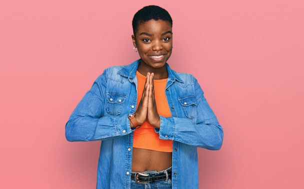 Afro-Amerikalı genç bir kadın günlük kıyafetler giyip ellerinin birliğiyle dua ediyor af diliyor kendinden emin bir şekilde gülümsüyor..  - Fotoğraf, Görsel