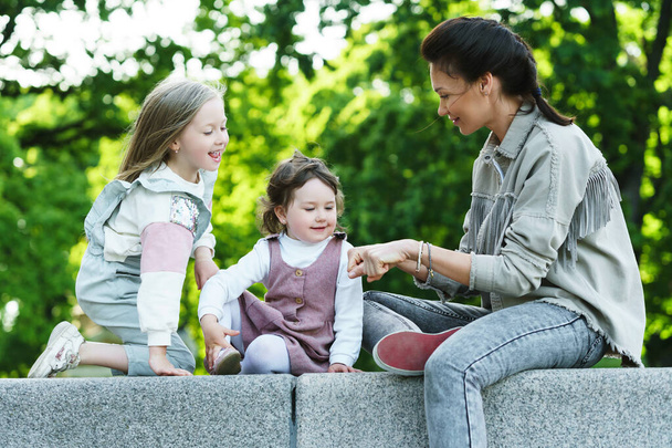Young happy mother and her two daughters sitting and playing in a city park - Фото, изображение