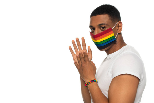 uomo africano americano in maschera medica e braccialetto di perline in colori lgbt guardando la fotocamera isolata su bianco - Foto, immagini